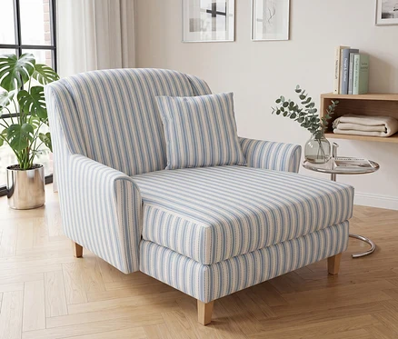 Ein hellblau-weiß gestreifter Chaiselongue mit passendem Kissen steht auf einem hellen Parkettboden in einem Wohnzimmer. Links sind ein großes Fenster mit weißen Vorhängen und eine Topfpflanze zu sehen, rechts ein Holzregal mit Büchern und einer Decke sowie ein Beistelltisch aus Glas mit einer Eukalyptusvase.