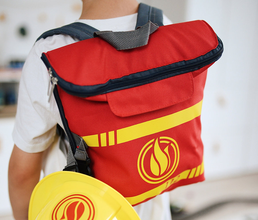 Ein Junge trägt einen roten small foot Feuerwehrrucksack und einen gelben Helm.