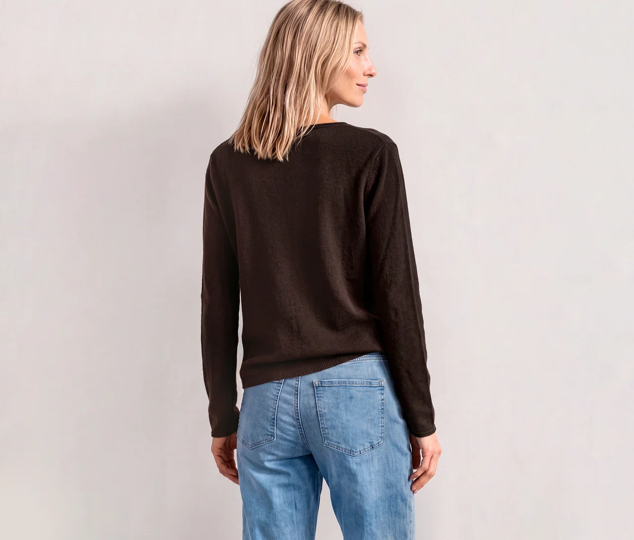 Frau von hinten in einem braunen Street One Strickpullover und hellblauen Jeans, blickt über ihre rechte Schulter.