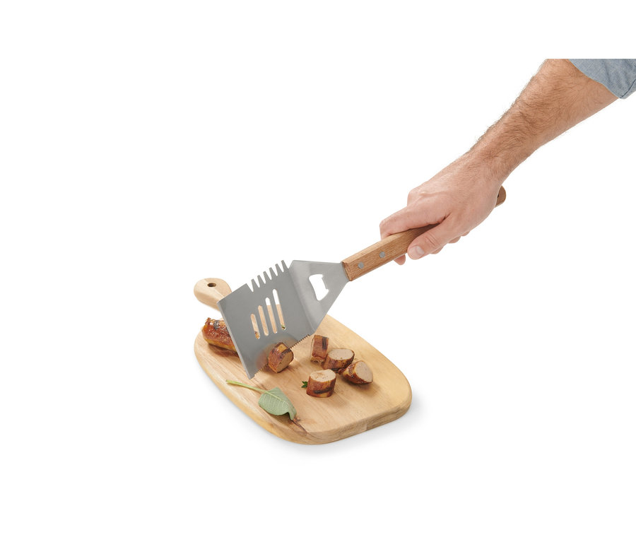 Eine Hand hält ein BBQ-Multitool über ein Holzbrett mit geschnittener Wurst.