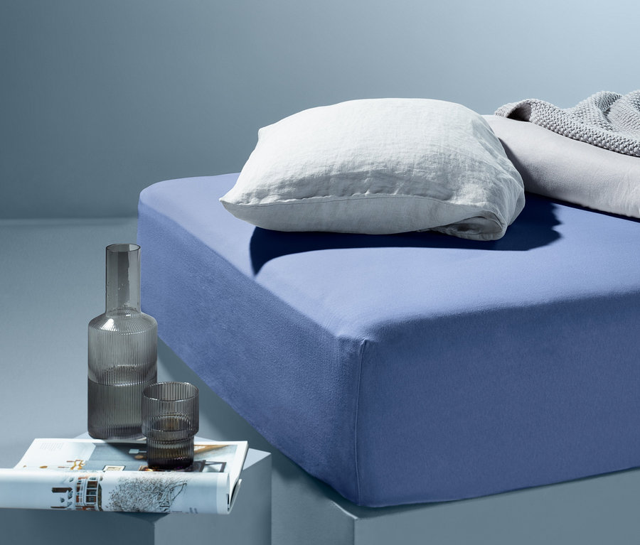 Blaues Jersey-Spannbettlaken in Normalgröße mit weißem Kissen und grauer Decke auf dem Bett.