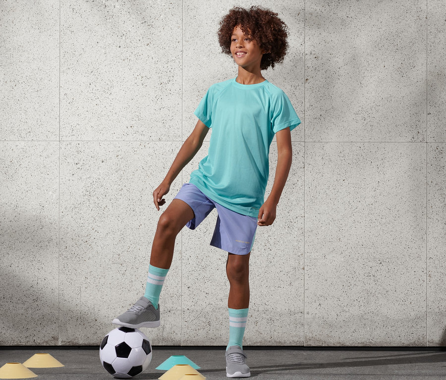 Ein Junge mit Afro steht mit einem Fuß auf einem Fußball. Er trägt 2 Funktionsshirts aus recyceltem Material in Türkis und lila Shorts.