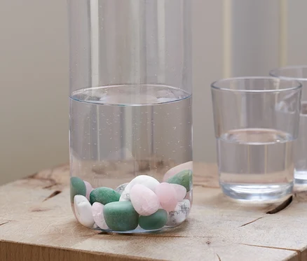 Eine klare Flasche mit Korkverschluss, teilweise gefüllt mit Wasser und Wasser-Steine, steht auf einem Holzblock neben zwei Gläsern Wasser.
