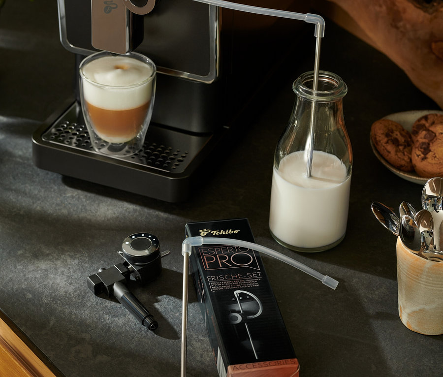 Ein anthrazitfarbener Tchibo »Esperto Pro« Kaffeevollautomat bereitet ein Getränk zu. Milch läuft durch einen Schlauch in eine Glasflasche. Daneben liegt ein Esperto Pro &amp; Esperto2 Milk Frische-Set.
