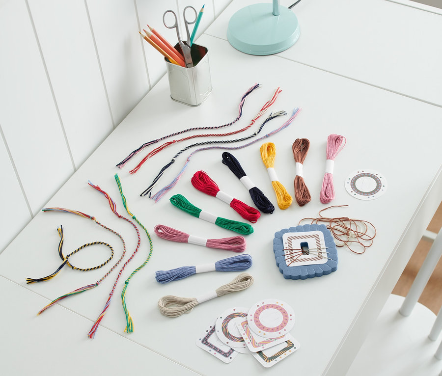 Auf einem weißen Tisch liegt ein Bastel-Set »Freundschaftsbänder« zusammen mit bunten Baumwollfäden, einer Schere und Buntstiften.