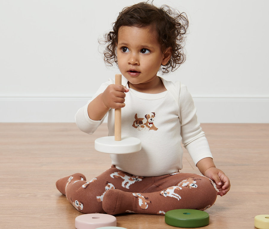 Ein Kind sitzt auf dem Boden und spielt mit einem Spielzeug. Es trägt eine 2 Paar Baby-Strumpfhose mit Hundemotiv.