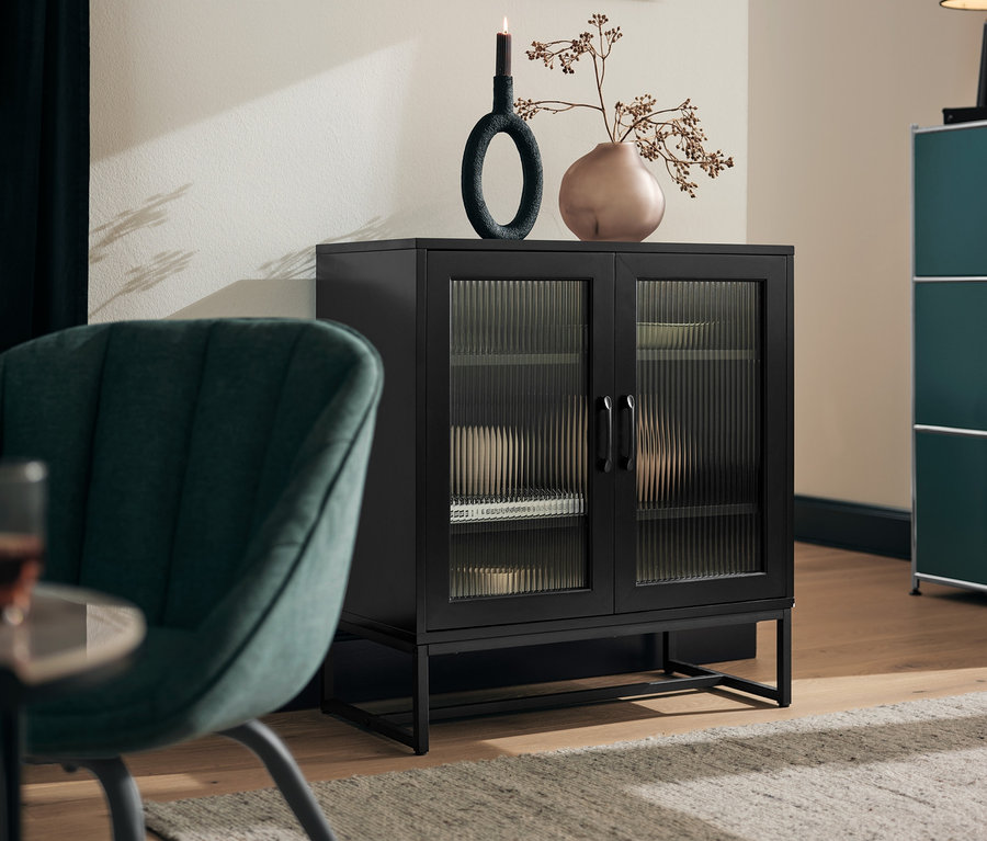 Ein grau-grüner, gepolsterter Stuhl steht neben einem schwarzen Sideboard mit Glastüren.