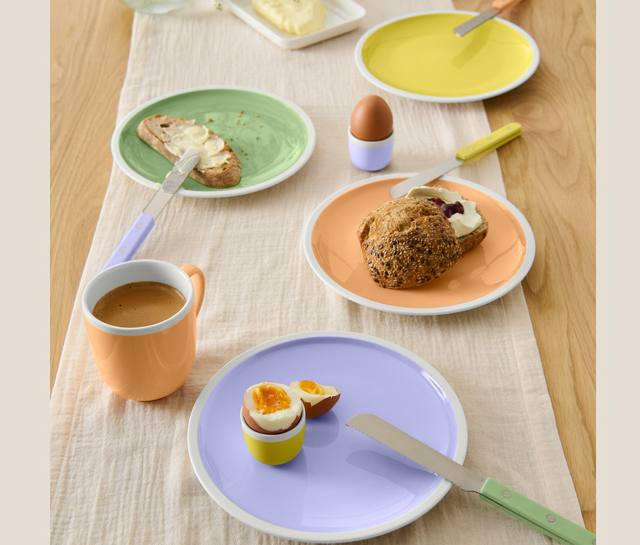 Auf dem Tisch liegen vier verschiedenfarbige Frühstücksteller mit Speisen. Auf den Tellern befinden sich Brot mit Butter, ein Ei und ein Brötchen mit Marmelade und Käse.