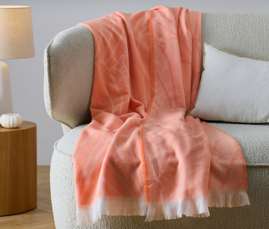 Eine orangefarbene Wende-Wohndecke mit Fransen liegt auf einem beigen Sofa neben einem weißen Kissen.