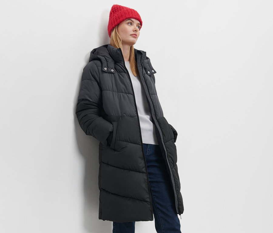 Frau posiert in Steppmantel mit Kapuze und roter Mütze.