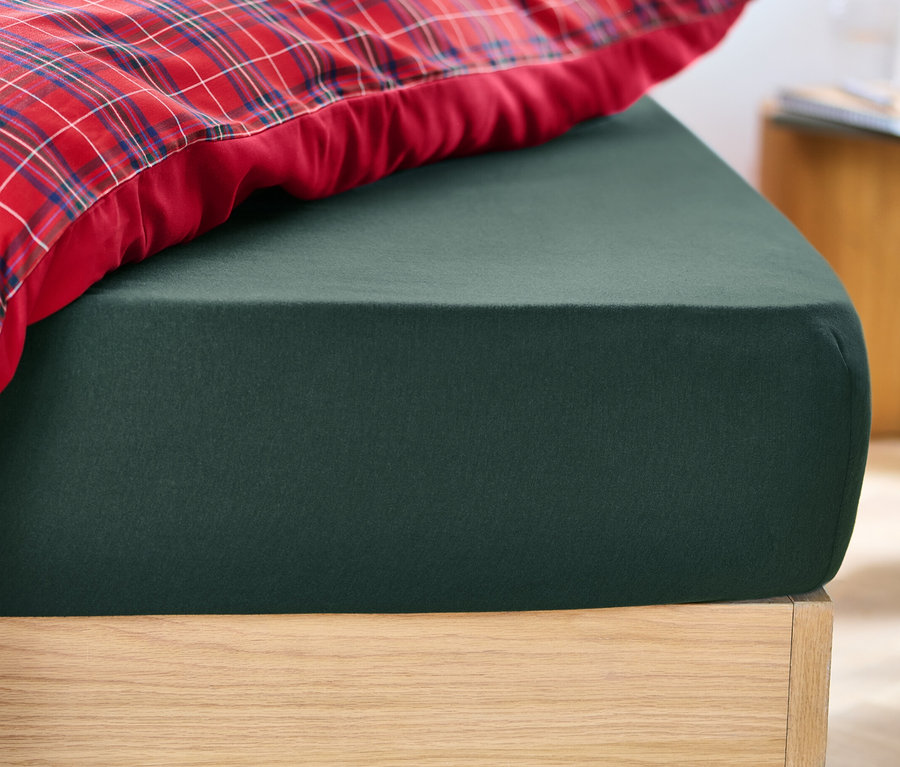 Grünes Biber-Spannbettlaken auf einem Bett neben einem rot karierten Kissen.
