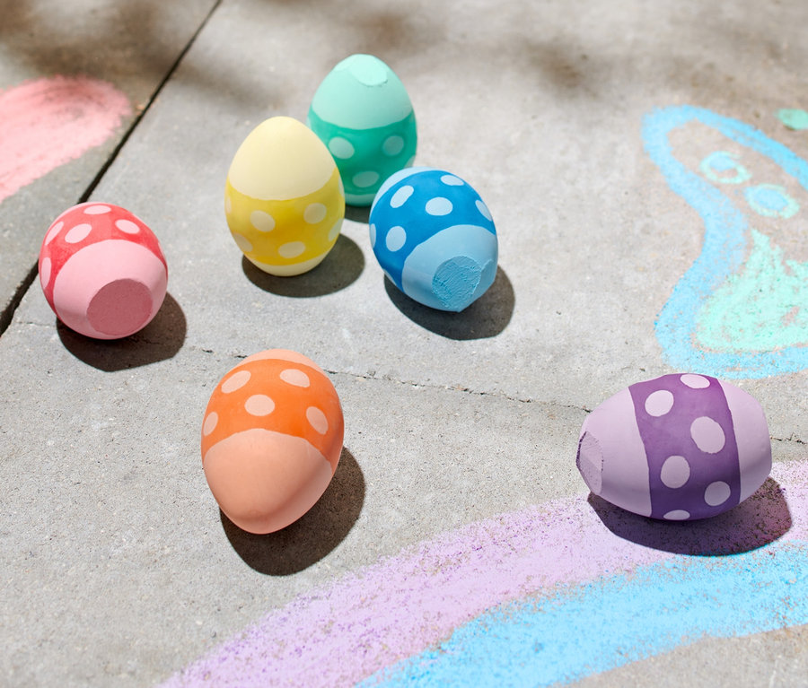 Sechs bunte Eier-förmige Straßenmalkreiden liegen auf einem Bürgersteig neben Kreidezeichnungen.