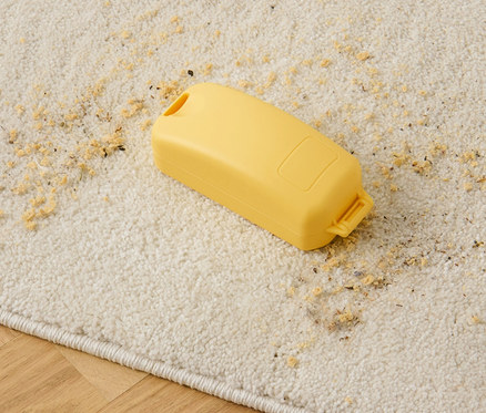 Eine gelbe Krümelbürste liegt auf einem beigen Teppich mit verstreuten Krümeln.
