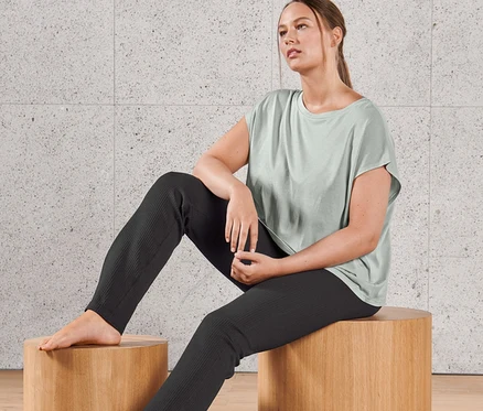 Frau sitzt auf einem Holzhocker und trägt ein Yogashirt und eine Seamless-Flared-Sporthose mit Schlitz.