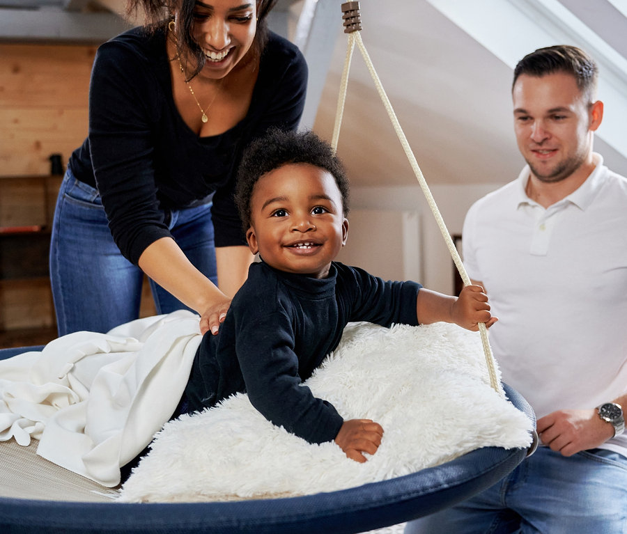 Ein Kind sitzt in einer schwarzen HUDORA Nestschaukel »Lounge« mit 90 cm Durchmesser, während die Mutter dahinter steht und der Vater daneben kniet.