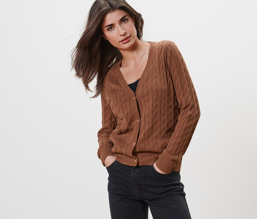 Frau posiert in Feinstrickcardigan mit Zopfmuster und dunkler Jeans.