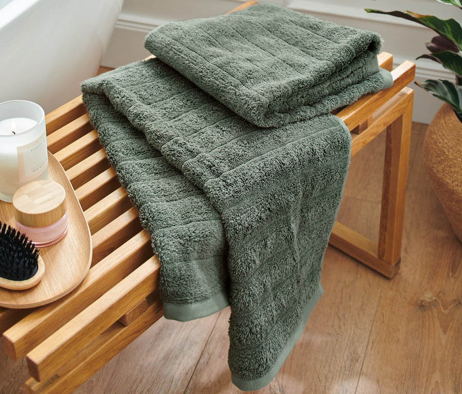 Zwei grüne Premium-Handtücher liegen gefaltet auf einer Holzbank neben einer Badewanne.