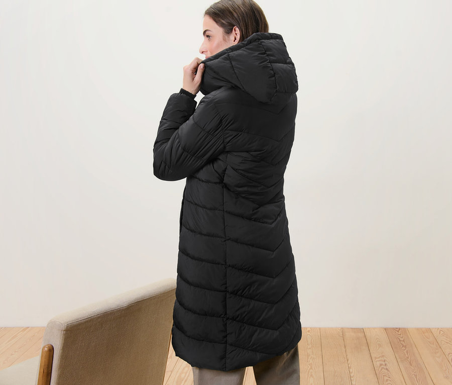 Frau justiert die Kapuze an einem schwarzen Steppmantel mit Kapuze.