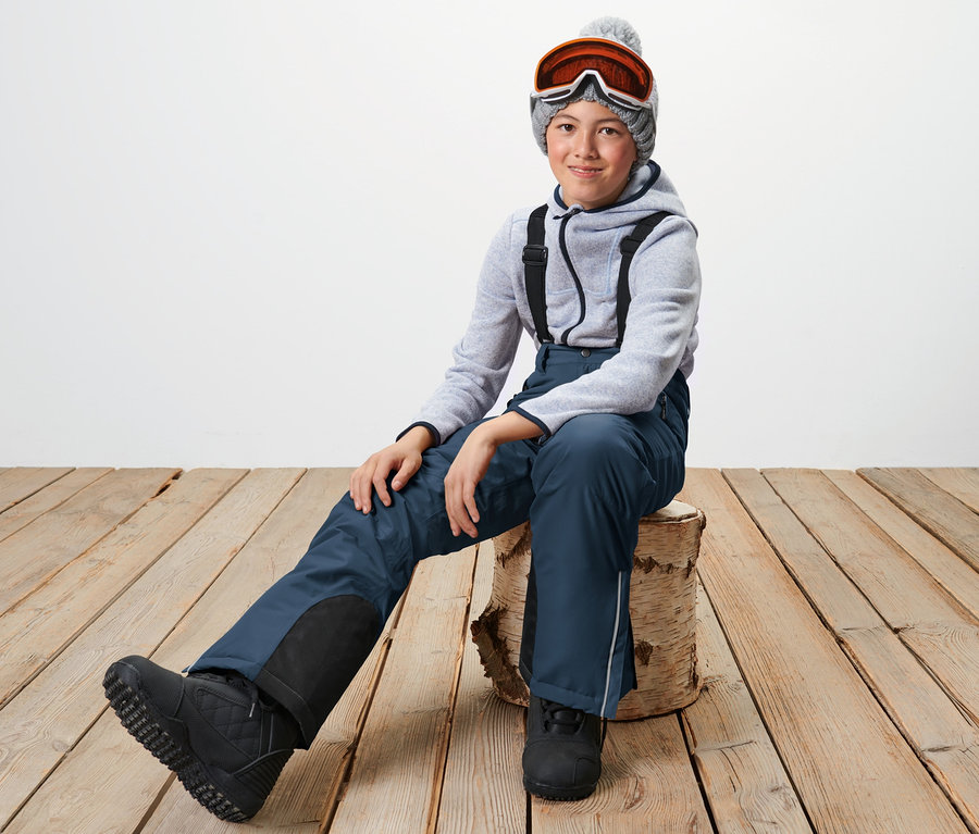 Ein Kind sitzt auf einem Holzschemel und trägt eine marineblaue Kinder-Skihose, einen Hoodie, eine Mütze und eine Skibrille.