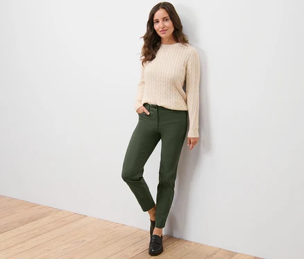 Frau posiert an einer Wand, trägt einen cremefarbenen Pullover und eine Bengalin-Hose.