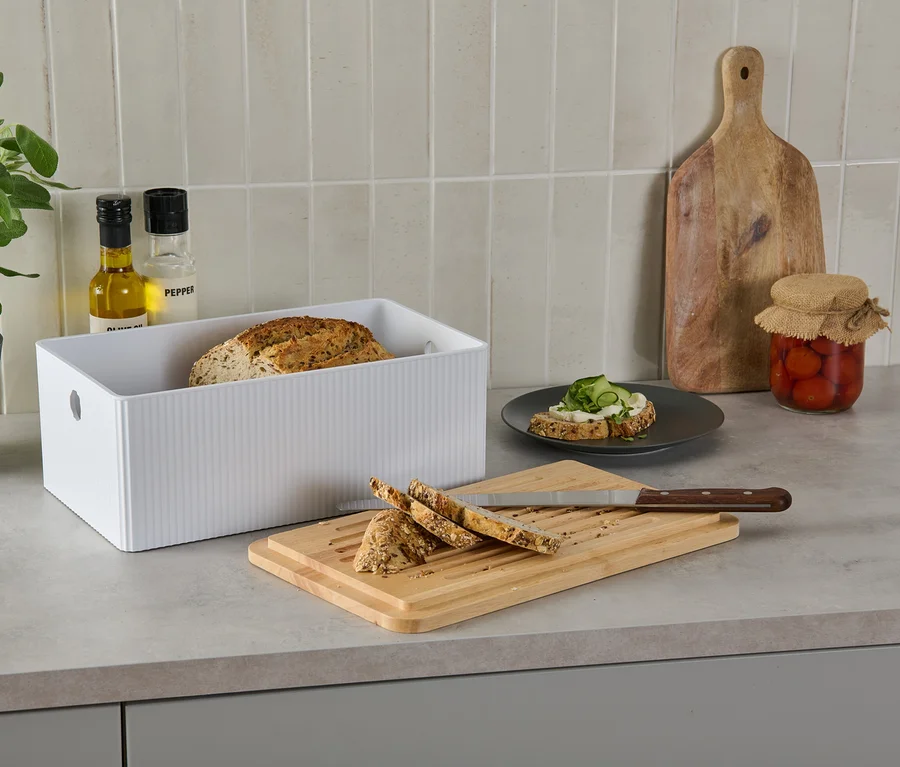 Brotkasten mit Schneidebrett auf einer Küchentheke neben Brot, einem belegten Brot, Öl, Pfeffer, einer Pflanze und eingelegten Tomaten.