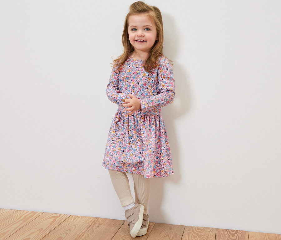 Lächelndes Mädchen trägt ein Kinder-Jerseykleid mit Blumenmuster und steht vor einer weißen Wand.