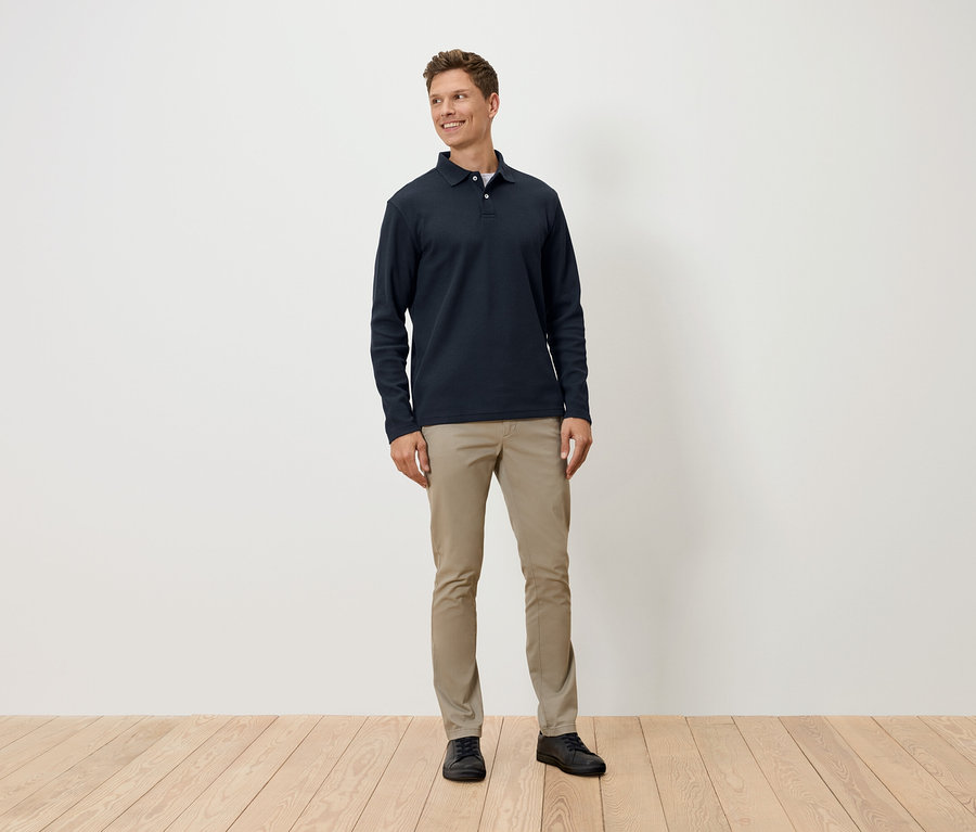 Mann steht und trägt ein dunkelblaues Langarm-Poloshirt, beige Hosen und schwarze Schuhe.