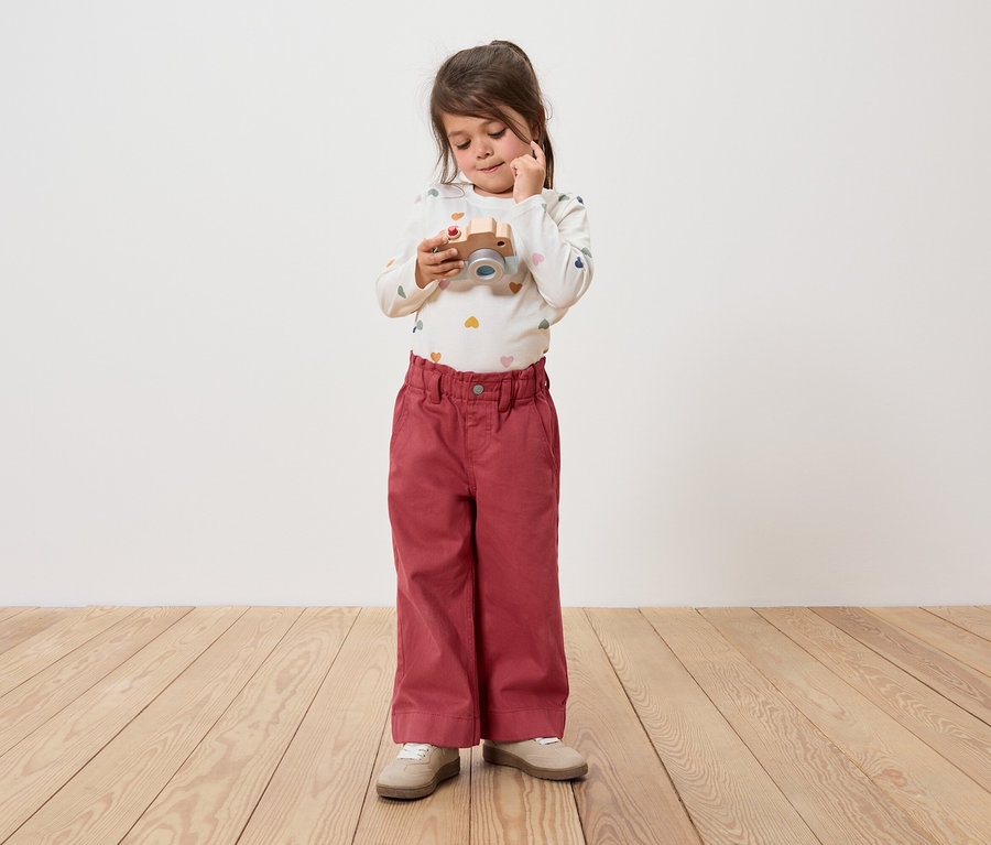 Mädchen hält eine Holzkamera und schaut sie an. Sie trägt eine Hose und ein langärmeliges Shirt.