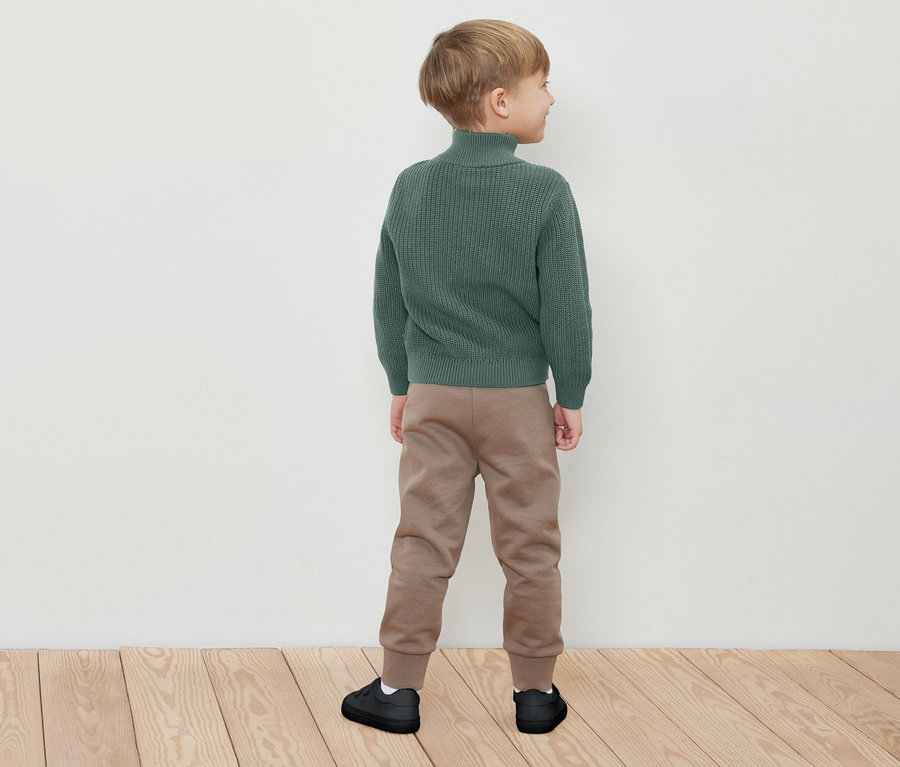 Ein Junge steht mit dem Rücken zur Kamera und trägt einen grünen Rollkragenpullover, braune Jogginghose und schwarze Turnschuhe.