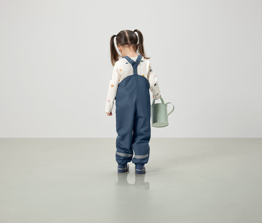 Mädchen in blauer Latzhose hält eine grüne Gießkanne.