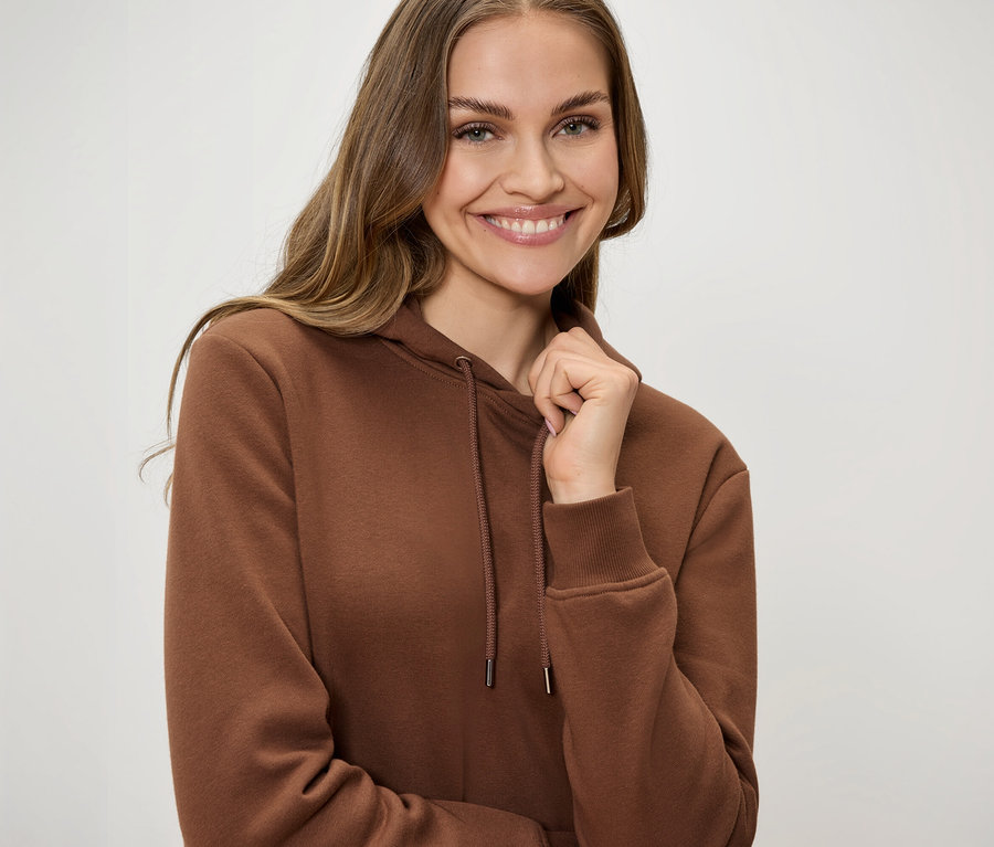 Lächelnde Frau in braunem Kapuzenpullover mit Kordelzug hält den Kragen mit einer Hand.