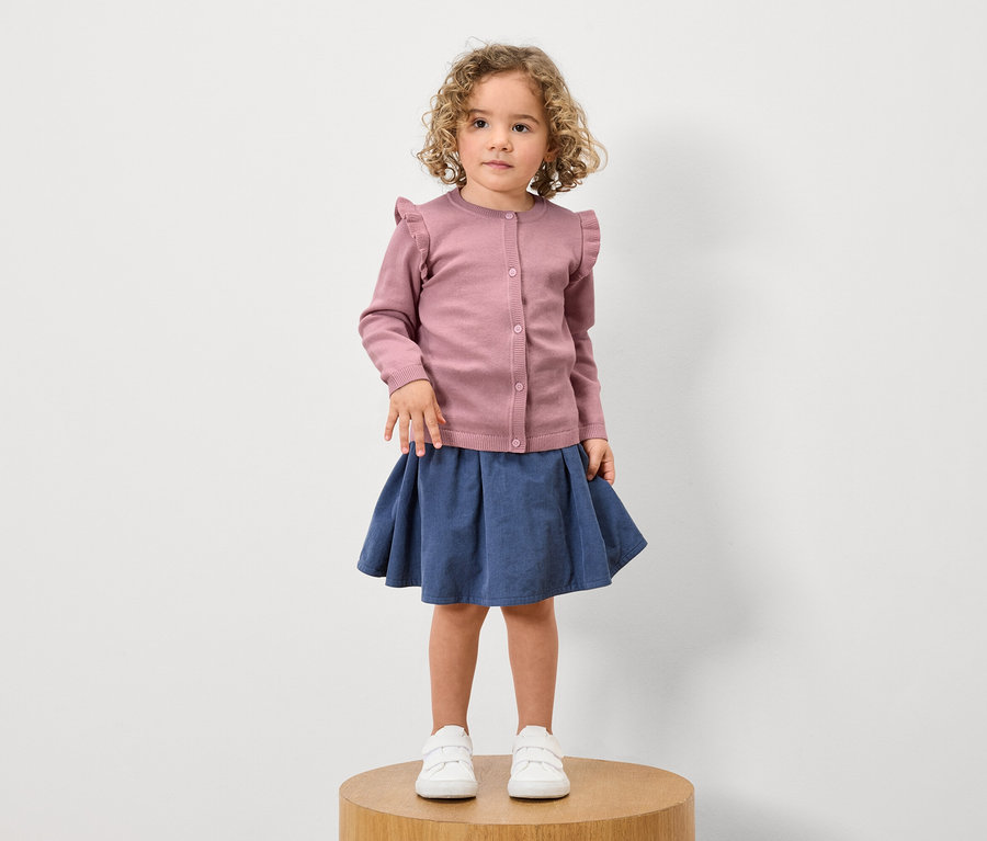 Mädchen mit lockigem Haar steht auf einem Holzsockel und trägt eine rosa Rüschenstrickjacke, einen blauen Rock und weiße Turnschuhe.
