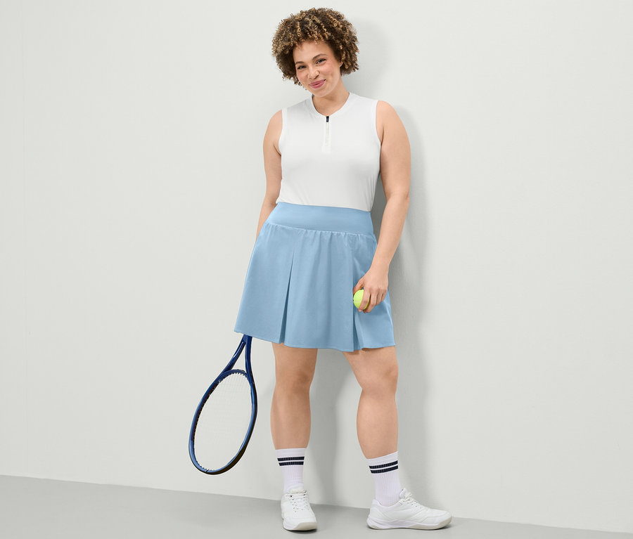Frau posiert in einem weißen Funktionstop und einem hellblauen Sportrock. Sie hält einen Tennisball und einen Tennisschläger.