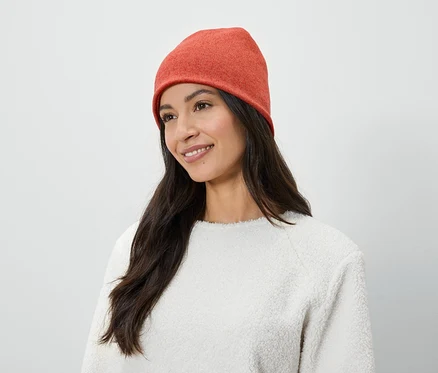 Frau mit langen braunen Haaren und einem Lächeln trägt eine orangefarbene Mütze und einen weißen Pullover.