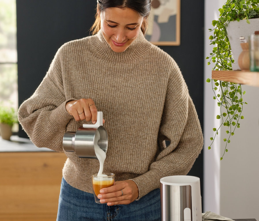 Frau gießt Milch aus einem Edelstahlbehälter in ein Glas neben einem schwarzen Induktions-Milchaufschäumer.