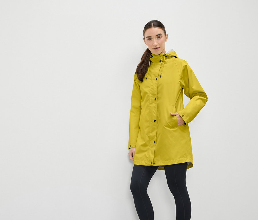 Eine Frau in einem gelben Regenmantel mit Kapuze und schwarzen Leggings steht vor einer weißen Wand.