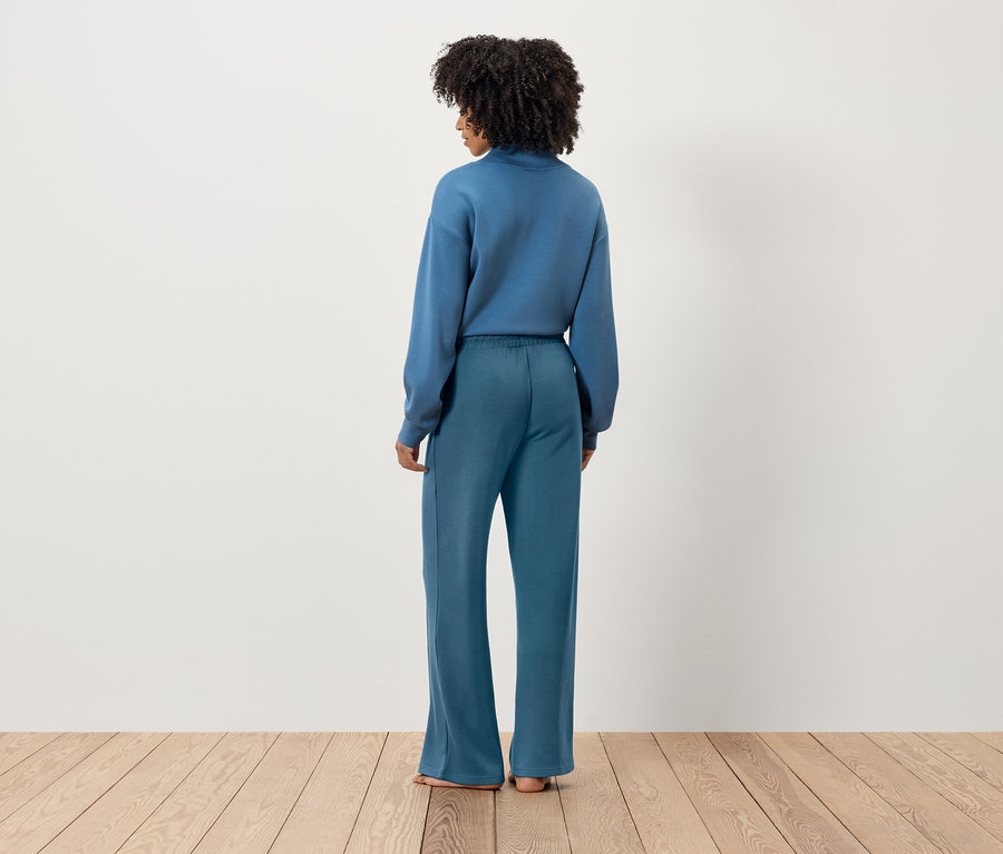 Rückansicht einer Frau mit dunklem lockigem Haar, die ein blaues Sweatshirt und eine weite Hose trägt.