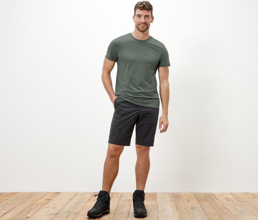 Mann trägt ein khaki Funktionsshirt, schwarze Shorts und schwarze Stiefel.
