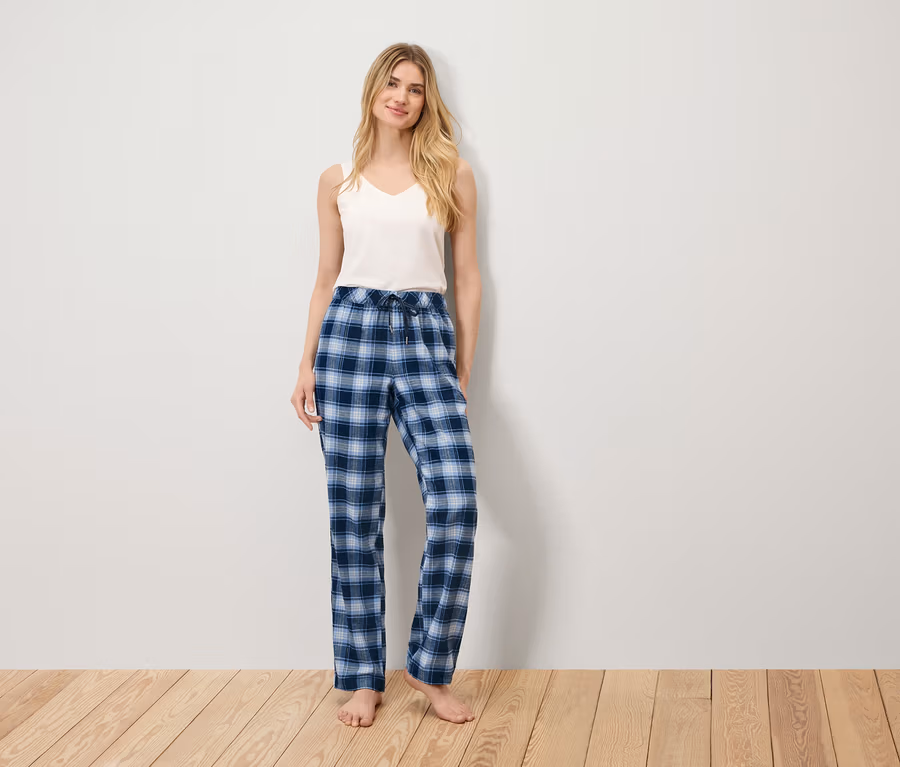 Eine Frau steht auf einem Holzboden und trägt ein weißes Tanktop und eine blau karierte Pyjamahose.