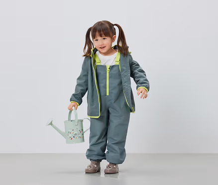 Mädchen in grauer Regenhose und Jacke hält hellgrüne Gießkanne.