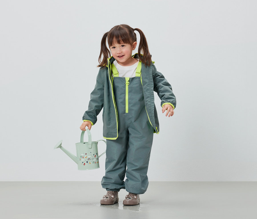 Mädchen in grauer Regenhose und Jacke hält hellgrüne Gießkanne.