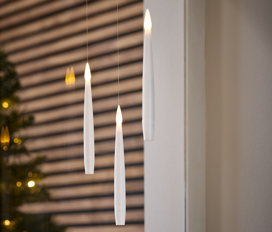 Drei weiße, elektronische Kerzen hängen an Fäden. Im Hintergrund befinden sich ein Weihnachtsbaum und eine Holzwand.