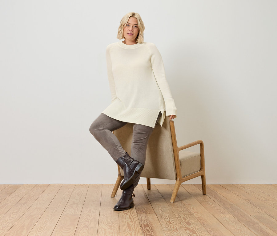 Blonde Frau posiert in einem langen, cremefarbenen Strickpullover und lehnt an einem Stuhl.