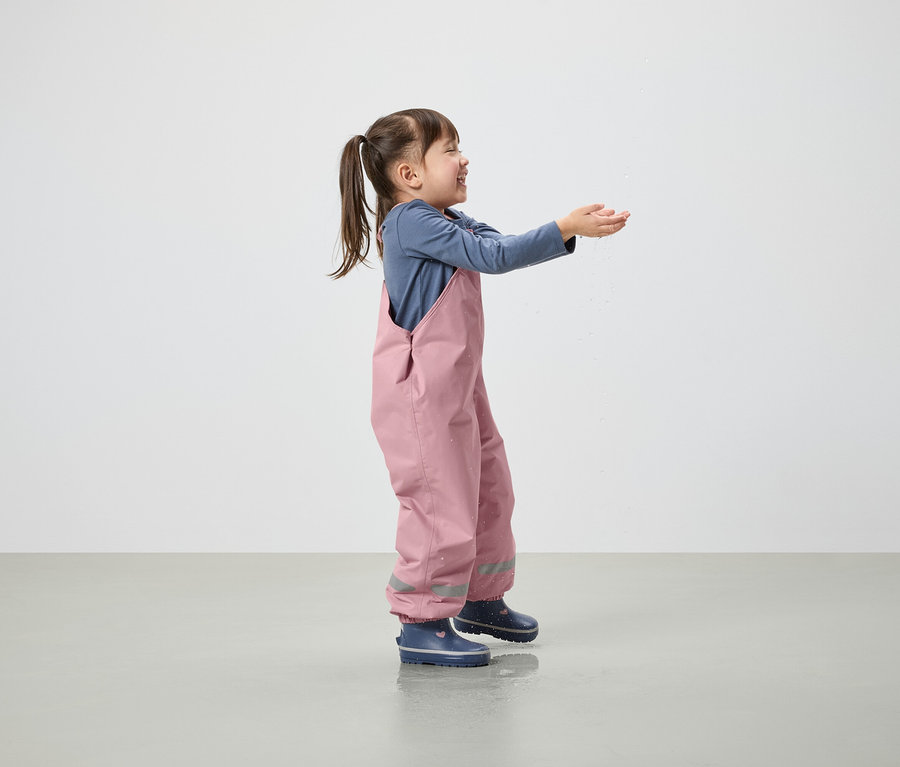 Seitenansicht eines lächelnden kleinen Mädchens mit Pferdeschwanz, das steht und die Hände ausstreckt. Sie trägt eine rosafarbene Latzhose und blaue Gummistiefel.