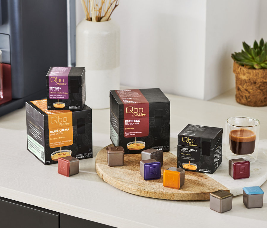 Auf einem Tisch stehen Qbo Espresso APANECA ANA Kapseln und eine Kaffeemaschine mit Espresso in einer Tasse.