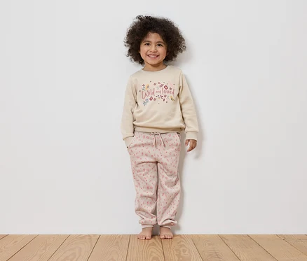 Ein lächelndes Mädchen mit lockigem Haar steht barfuß vor einer weißen Wand und trägt einen beigen Pullover mit der Aufschrift "Wild and loved" und rosafarbene Hosen.