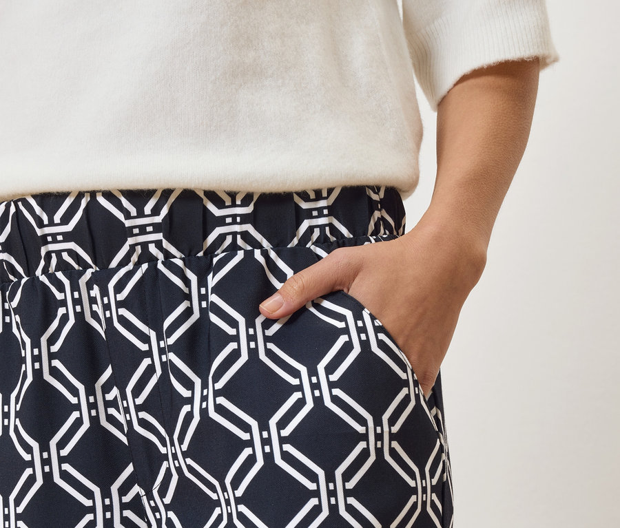 Nahaufnahme einer Person, die eine marineblaue Hose mit weißem geometrischen Muster und einen weißen Pullover trägt, mit der Hand in der Hosentasche.