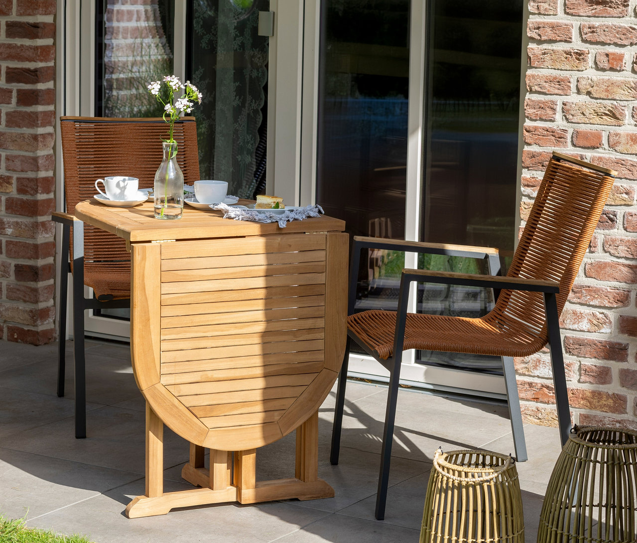 Auf einer Terrasse stehen zwei Stühle mit braunem Geflecht an einem Holztisch mit Geschirr und Blumen.
