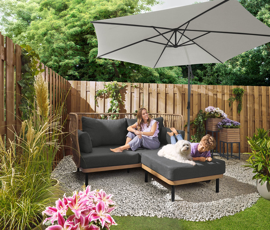 Eine Frau sitzt auf einem grauen Sofa unter einem Leco Ampelschirm Ø 300 cm mit Schirmständer und Abdeckhaube. Ein Kind liegt mit einem Hund auf einem Ottoman. Im Hintergrund ist ein Leco Plattenset für Kreuzständer zu sehen.