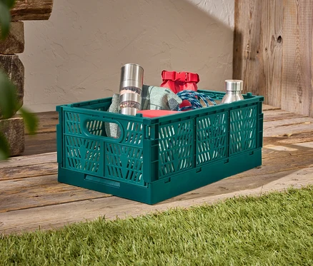 Eine grüne Kunststoffbox steht auf einem Holzboden, gefüllt mit einer Edelstahl-Pfeffermühle, einer roten Tasche, einem blauen Seil und einer Edelstahlflasche.